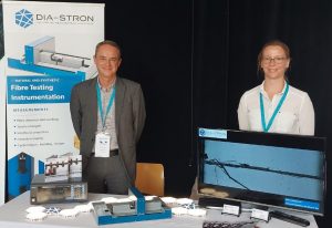 Managing Director Yann Leray and European Territory Manager Maud Dumas on Dia-Stron's stand at the EPNOE Conference in Nantes. The fibra.stress instrument is in the centre of the table-top display, alongside brochures and a screen showing close-up footage of a fibre breaking. Behind the stand is a pop-up banner showing the fibra.stress under the Dia-Stron logo, with the caption "Natural and Synthetic Fibre Testing Instrumentation"