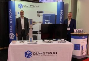 Dia-Stron's Head of Marketing and Sales Dr Richard Blackwell-Whitehead and Managing Director Yann Leray on Dia-Stron's booth at NYSCC Suppliers' Day. A fibra.one with combing accessory is set-up on a covered table with brochures laid out, with a large branded banner at the back of the stand showing the MTT690 tensile tester. Yann stands next to a branded podium printed with photos of the fibra.one